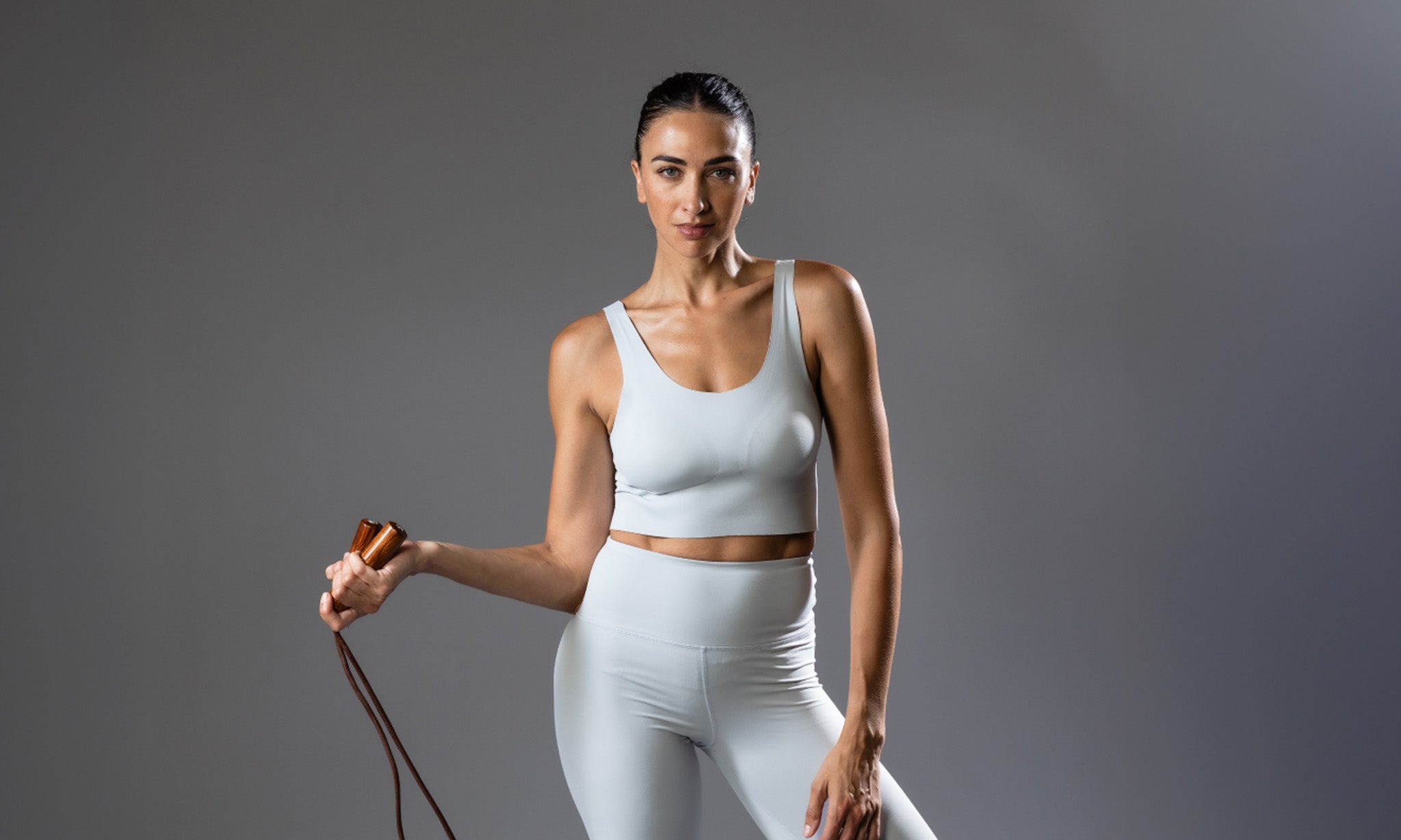 Woman in a light gray athletic outfit holding a jump rope against a dark background